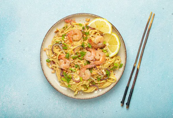 Karides, fıstık, yeşil soğan, limon dilimleri ile süslenmiş Tayland erişteleri ve açık mavi arka planda servis edilen çubuklarla Tayland mutfağı.
