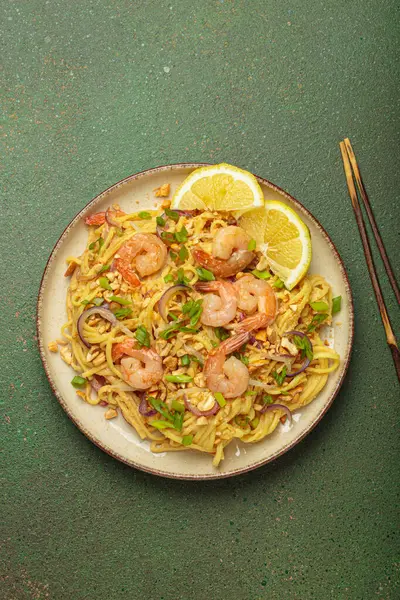 Karides, fıstık, yeşil soğan, limon dilimleri ile süslenmiş Tayland erişteleri ve kırsal yeşil arka planda servis edilen çubuklarla Tayland mutfağı.