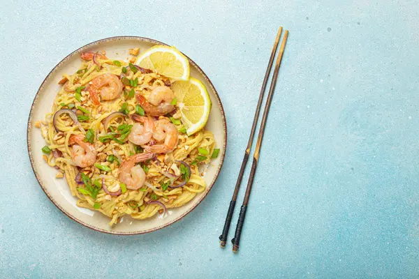 Karides, fıstık, yeşil soğan, limon dilimleri ile süslenmiş Tayland erişteleri ve açık mavi arka planda çubuklarla servis edilmiş Tayland mutfağı, kopyalama alanı.