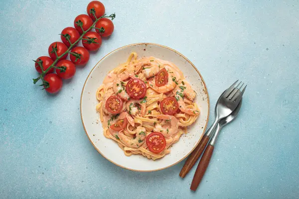 Taze maydanozla süslenmiş karides ve kiraz domatesli kaymak soslu deniz ürünleri linguine makarna, açık mavi arka planda servis edilir.