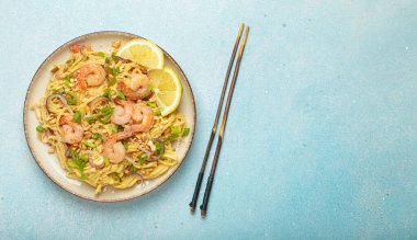 Karides, fıstık, yeşil soğan, limon dilimleri ile süslenmiş Tayland erişteleri ve açık mavi arka planda çubuklarla servis edilmiş Tayland mutfağı, kopyalama alanı.