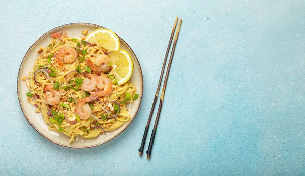 Karides, fıstık, yeşil soğan, limon dilimleri ile süslenmiş Tayland erişteleri ve açık mavi arka planda çubuklarla servis edilmiş Tayland mutfağı, kopyalama alanı.