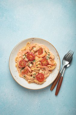Taze maydanozla süslenmiş karides ve kiraz domatesli kaymak soslu deniz ürünleri linguine makarna, açık mavi arka planda servis edilir.