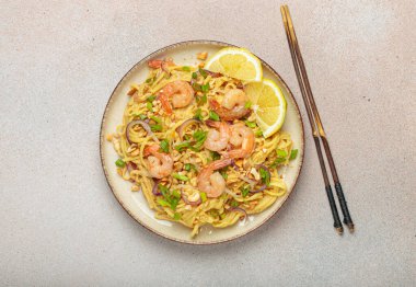 Karides, fıstık, yeşil soğan, limon dilimleri ile süslenmiş Tayland eriştesi bej arka planda çubuklarla servis edilmiş Tayland mutfağı.