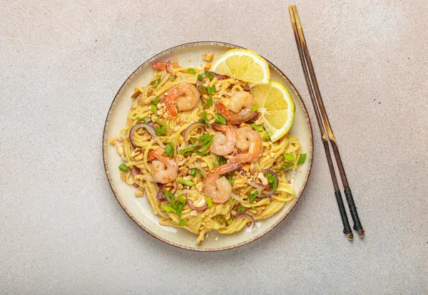 Karides, fıstık, yeşil soğan, limon dilimleri ile süslenmiş Tayland eriştesi bej arka planda çubuklarla servis edilmiş Tayland mutfağı.