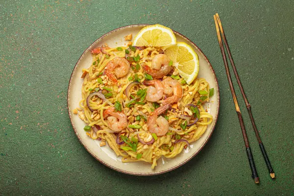 Karides, fıstık, yeşil soğan, limon dilimleri ile süslenmiş Tayland erişteleri ve kırsal yeşil arka planda servis edilen çubuklarla Tayland mutfağı.