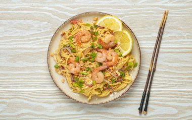 Karides, fıstık, yeşil soğan, limon dilimleri ile süslenmiş Tayland erişteleri ve beyaz ahşap bir masada servis edilen çubuklarla Tayland mutfağı.