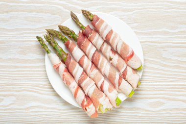 Pastırma şeritlerine sarılı lezzetli kuşkonmaz, renkli biberlerle tatlandırılmış, beyaz bir tabakta sunulmuş, pişirilmeye hazır.