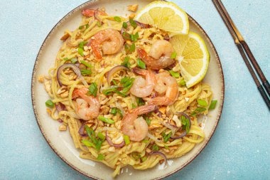 Karides, fıstık, yeşil soğan, limon dilimleri ile süslenmiş Tayland erişteleri ve açık mavi arka planda servis edilen çubuklarla Tayland mutfağı.