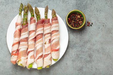 Pastırma şeritlerine sarılı lezzetli kuşkonmaz, renkli biberlerle tatlandırılmış, beyaz bir tabakta sunulmuş, pişirilmeye hazır.