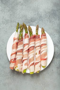 Pastırma şeritlerine sarılı lezzetli kuşkonmaz, renkli biberlerle tatlandırılmış, beyaz bir tabakta sunulmuş, pişirilmeye hazır.