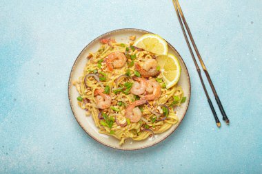 Karides, fıstık, yeşil soğan, limon dilimleri ile süslenmiş Tayland erişteleri ve açık mavi arka planda servis edilen çubuklarla Tayland mutfağı.