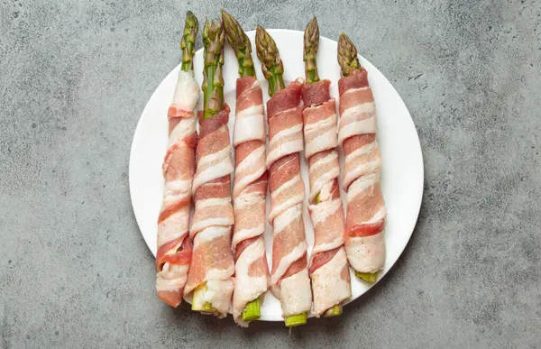 Pastırma şeritlerine sarılı lezzetli kuşkonmaz, renkli biberlerle tatlandırılmış, beyaz bir tabakta sunulmuş, pişirilmeye hazır.