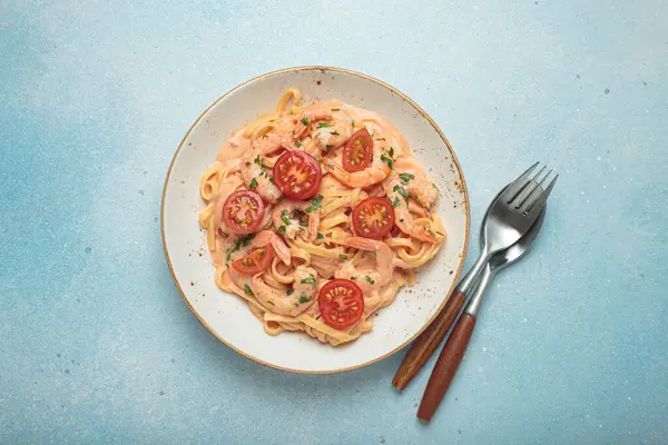 Taze maydanozla süslenmiş karides ve kiraz domatesli kaymak soslu deniz ürünleri linguine makarna, açık mavi arka planda servis edilir.