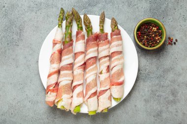 Pastırma şeritlerine sarılı lezzetli kuşkonmaz, renkli biberlerle tatlandırılmış, beyaz bir tabakta sunulmuş, pişirilmeye hazır.