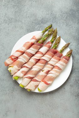 Pastırma şeritlerine sarılı lezzetli kuşkonmaz, renkli biberlerle tatlandırılmış, beyaz bir tabakta sunulmuş, pişirilmeye hazır.