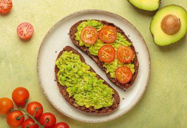 İki dilim tam çavdar ekmeği üzerine püre avokado, yeşil bezelye, kiraz domatesleri ve susam tohumları, sağlıklı ve canlı bir kahvaltı veya aperatif seçeneği konur.