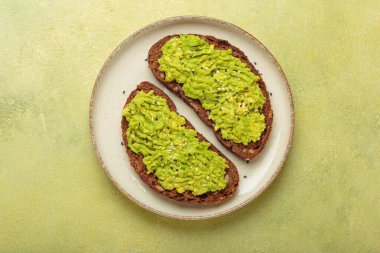 Susam tohumlu ezilmiş avokado iki dilim kızarmış çavdar ekmeğine yayılır, yeşil bir arkaplanda servis edilir, sağlıklı ve lezzetli bir kahvaltı veya atıştırmalık