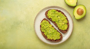 İki dilim kızarmış çavdar ekmeği üzerine püre avokado ve susam tohumu serpiştirilir, bir tabakta servis edilir, yarım avokado, sağlıklı ve enerjik bir kahvaltı veya atıştırmalık seçeneğinin yanında.