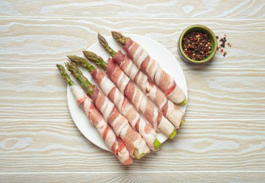 Pastırma şeritlerine sarılı lezzetli kuşkonmaz, renkli biberlerle tatlandırılmış, beyaz bir tabakta sunulmuş, pişirilmeye hazır.