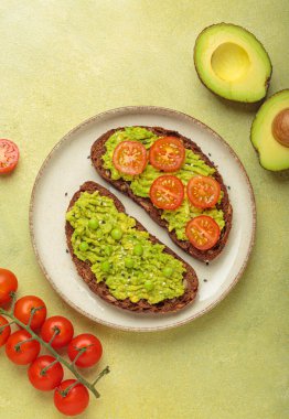 İki dilim tam çavdar ekmeği üzerine püre avokado, yeşil bezelye, kiraz domatesleri ve susam tohumları, sağlıklı ve canlı bir kahvaltı veya aperatif seçeneği konur.
