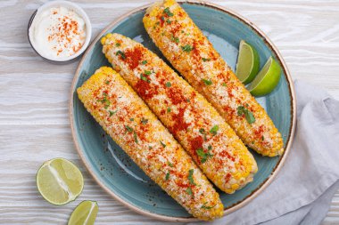 Üç ızgara mısır koçanı baharat, mayonez ve cotija peyniri ile kaplı limon dilimleri ve acı biber tozlu krema ile servis edilir.