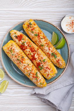 Üç ızgara mısır koçanı baharat, mayonez ve cotija peyniri ile kaplı limon dilimleri ve acı biber tozlu krema ile servis edilir.