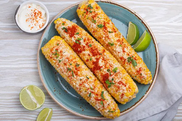 Üç ızgara mısır koçanı baharat, mayonez ve cotija peyniri ile kaplı limon dilimleri ve acı biber tozlu krema ile servis edilir.