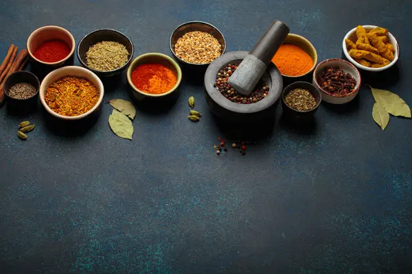 Aromatik baharat ve otlarla dolu renkli kaseler koyu bir arka planda, harç ve tokmakla bir sınır oluşturuyor. Gurme mutfağı için aromalı malzemelerin üst görünümü, kopya alanı sunar
