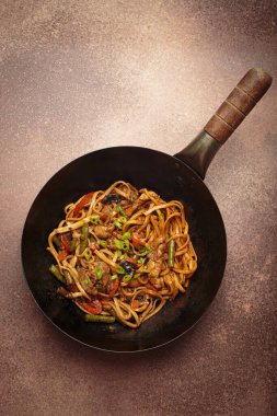 Tavada kızarmış tavuk ve sebzeli erişte, tepesinde ahşap çubuklar, kırsal kahverengi arka planda üst manzara.