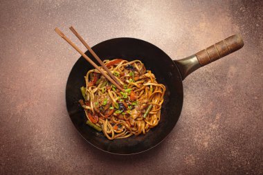 Tavada kızarmış tavuk ve sebzeli erişte, tepesinde ahşap çubuklar, kırsal kahverengi arka planda üst manzara.