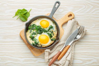 Sağlıklı ıspanaklı omlet, çırpılmış demir bir tavada iki sahanda yumurta, ahşap bir masada çatal bıçak ve peçeteyle servis edilir, besleyici bir kahvaltı veya brunch için mükemmeldir.