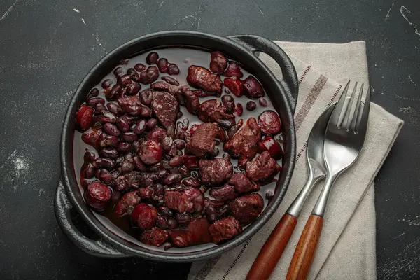 Feijoada, dökme demir bir tencerede geleneksel Brezilya güveci, keten peçetede çatal ve kaşıkla servis edilmeye hazır.