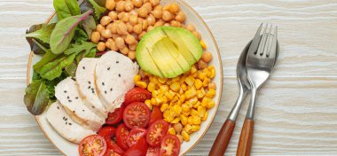 Kızarmış tavuk göğsü, avokado, nohut, mısır, domates ve karışık yeşillikler, yukarıdan beyaz ahşap bir masa, dengeli ve besleyici bir yemek.