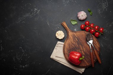 Kiraz domatesleri, kırmızı dolma biber ve taze fesleğen yaprakları ahşap bir kereste kesme tahtasının yanında koyu dokulu bir arka planda duruyor.