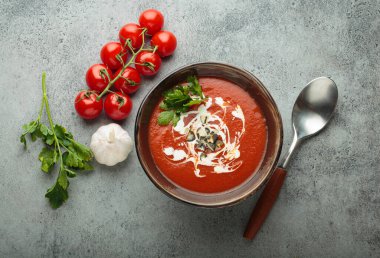 Krema, tohum ve otlarla dolu bir kasede taze domates çorbası servis edilir, yanında da kaşık ve peçeteyle birlikte kiraz domatesleri, sarımsak ve maydanoz bulunur.