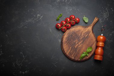 Kiraz domatesleri, sarımsak ve taze fesleğen yaprakları ahşap bir kereste kesme tahtasının üzerinde, ahşap bir biber değirmeninin yanında, koyu renkli bir arka plan, kopyalama alanı.