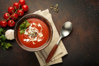 Krema, tohum ve otlarla dolu bir kasede taze domates çorbası servis edilir. Yanında da kaşık ve peçeteyle kahverengi bir zemin üzerinde domates, sarımsak ve maydanoz bulunur.