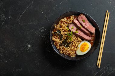 Bir kase erişte çorbası dilimlenmiş sığır eti, haşlanmış yumurta, deniz yosunu ve yeşil soğan, koyu arkaplanda çubuklarla servis ediliyor, Asya usulü çorba üstü görünümü, kopyalama alanı.