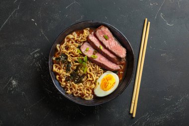 Bir kase erişte çorbası dilimlenmiş sığır eti, haşlanmış yumurta, yosun ve yeşil soğan, koyu arkaplanda çubuklarla servis ediliyor, Asya usulü çorba üstü manzaralı.