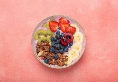 Dilimlenmiş kivi, çilek, yabanmersini, kiraz, muz ve granola ile kaplı canlı ve besleyici acai kasesi, pembe arka planda renkli ve sağlıklı bir yemek veya kahvaltı