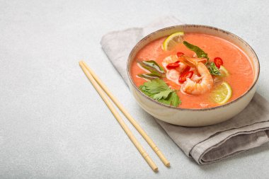 Taze karides, limon kamaları, şifalı otlar ve kırmızı biberlerle süslenmiş ve yanında keten peçetede yemek çubukları bulunan Tayland 'ın popüler bir yemeği olan kremalı Tom yum çorbası bir kâse içinde servis edilir.