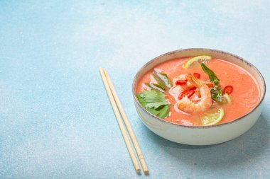 Taze karides, limon kamaları, şifalı otlar ve kırmızı biberlerle süslenmiş ve yanında keten peçetede yemek çubukları bulunan Tayland 'ın popüler bir yemeği olan kremalı Tom yum çorbası bir kâse içinde servis edilir.