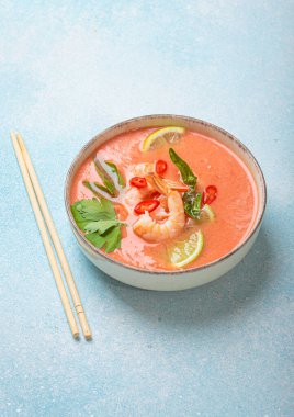 Taze karides, limon kamaları, şifalı otlar ve biberlerle süslenmiş ve yanında keten peçetede yemek çubukları bulunan Tayland 'ın popüler bir yemeği olan kremalı Tom yum çorbası bir kasede servis edilir.