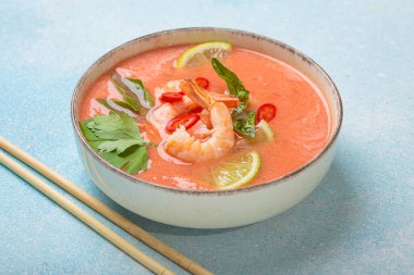 Taze karides, limon kamaları, şifalı otlar ve biberlerle süslenmiş ve yanında keten peçetede yemek çubukları bulunan Tayland 'ın popüler bir yemeği olan kremalı Tom yum çorbası bir kasede servis edilir.