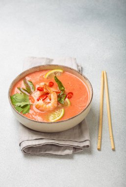 Taze karides, limon kamaları, şifalı otlar ve biberlerle süslenmiş ve yanında keten peçetede yemek çubukları bulunan Tayland 'ın popüler bir yemeği olan kremalı Tom yum çorbası bir kasede servis edilir.