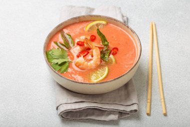 Taze karides, limon kamaları, şifalı otlar ve biberlerle süslenmiş ve yanında keten peçetede yemek çubukları bulunan Tayland 'ın popüler bir yemeği olan kremalı Tom yum çorbası bir kasede servis edilir.