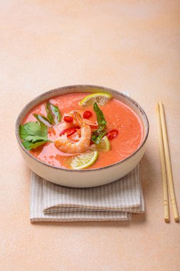 Taze karides, limon kamaları, şifalı otlar ve biberlerle süslenmiş ve yanında keten peçetede yemek çubukları bulunan Tayland 'ın popüler bir yemeği olan kremalı Tom yum çorbası bir kasede servis edilir.