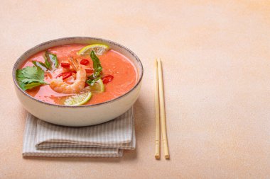 Taze karides, limon kamaları, şifalı otlar ve kırmızı biberlerle süslenmiş ve yanında keten peçetede yemek çubukları bulunan Tayland 'ın popüler bir yemeği olan kremalı Tom yum çorbası bir kâse içinde servis edilir.