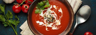 Taze domates çorbası krema, tohum ve otlarla dolu bir kasede servis edilir, yanında da mavi bir zemin üzerinde kaşık ve peçeteyle kiraz domatesleri, sarımsak ve maydanoz bulunur.